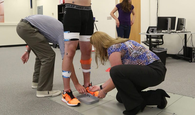 Male standing on force plates with two people placing retroflective markers on his lower limbs.