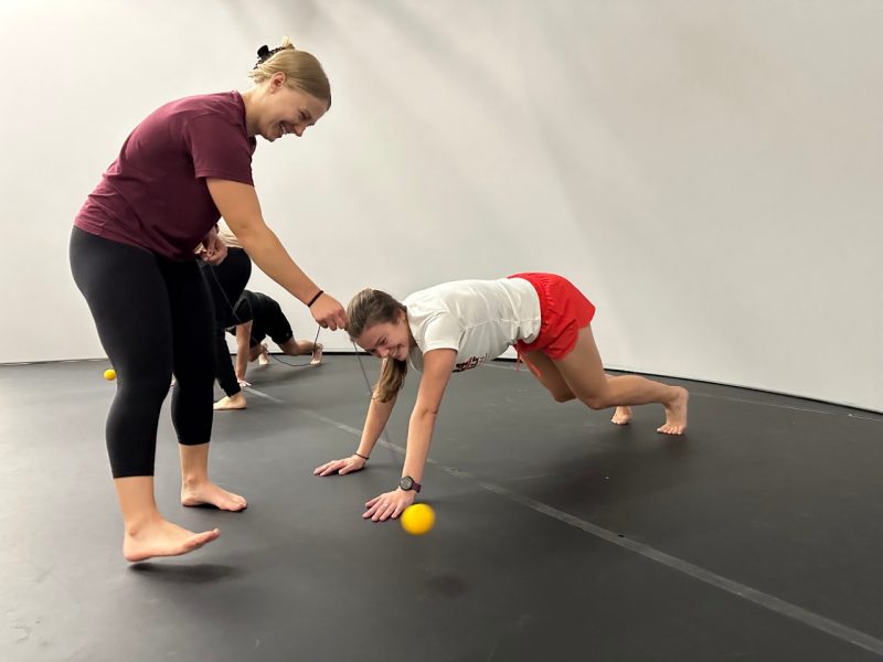 A woman wearing a maroon shirt and black leggings swining a yellow ball on a string near a woman wearing a white shirt and red shorts in a plank on the ground.
